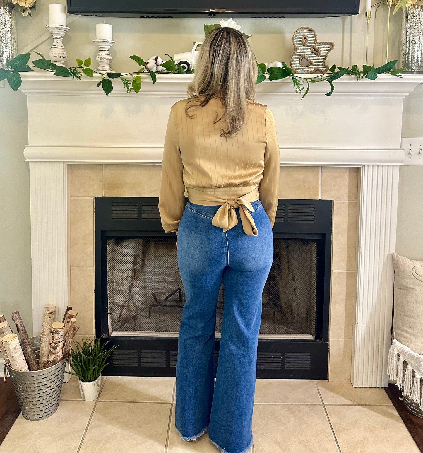 Woman standing in front of a fireplace wearing a beige top and blue jeans.