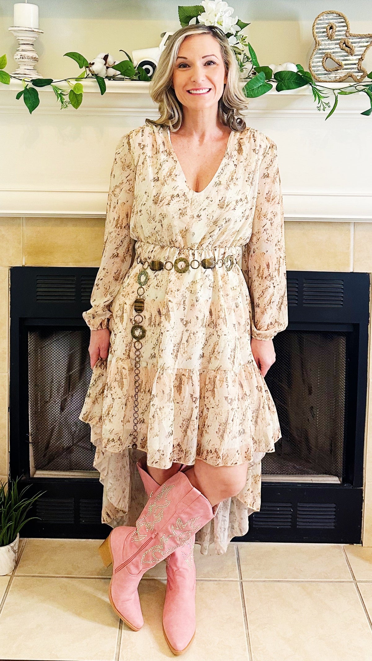 A woman stands in front of a fireplace wearing a long-sleeve, tiered cream floral dress with a decorative belt and pink cowboy boots.