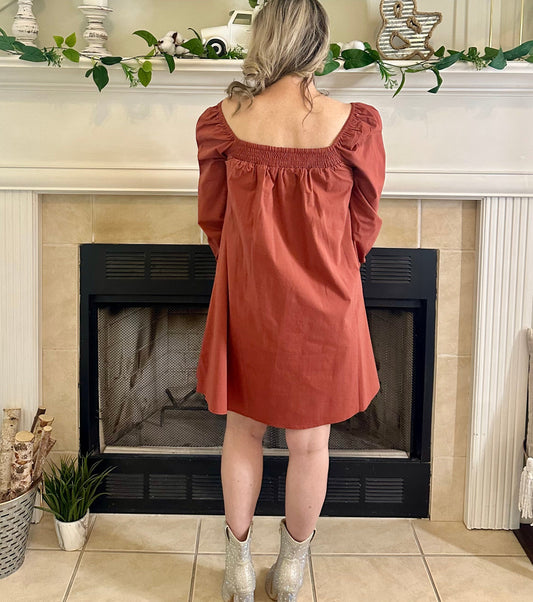 Woman in a rust-colored dress standing in front of a fireplace with decorative elements.
