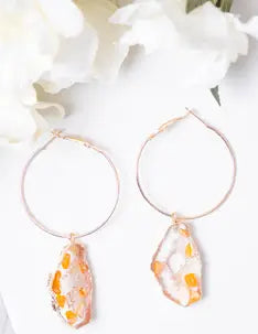 Gold hoop earrings with orange teardrop-shaped stones on a white background with flowers.