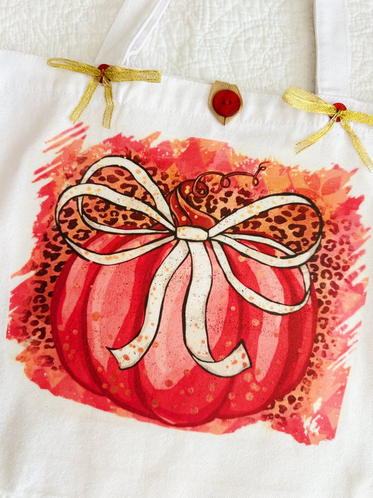 A wwhite canvas tote bag featuring a printed autumn design of a red pumpkin with a leopard-print bow and a red watercolor background, resting on a pink patterned surface.