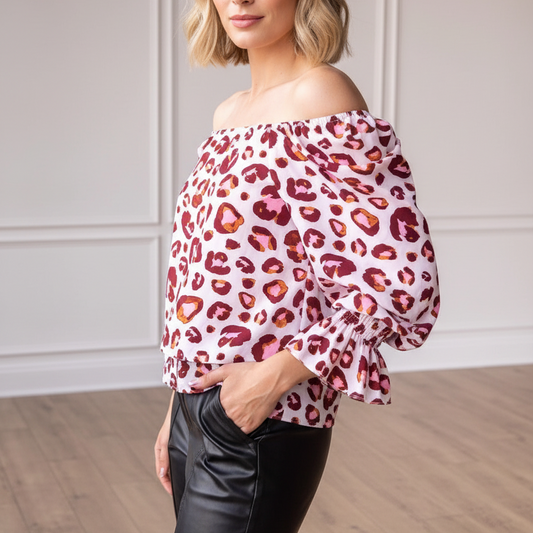 Woman wearing a leopard print off-shoulder top and black pants in a room with wooden floor and white walls.