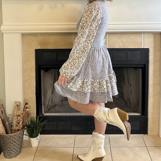 Woman in a light blue dress and white boots standing in front of a fireplace with decorative items.