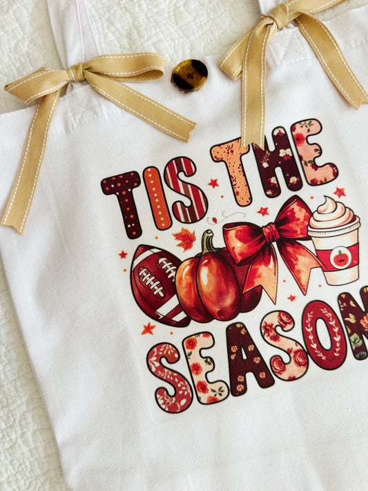 A white canvas tote bag with a gold ribbon handle bow, featuring a fall-themed graphic that reads 'TIS THE SEASON' in autumn patterned letters, alongside pumpkins, a football, a bow, and a hot cocoa cup.