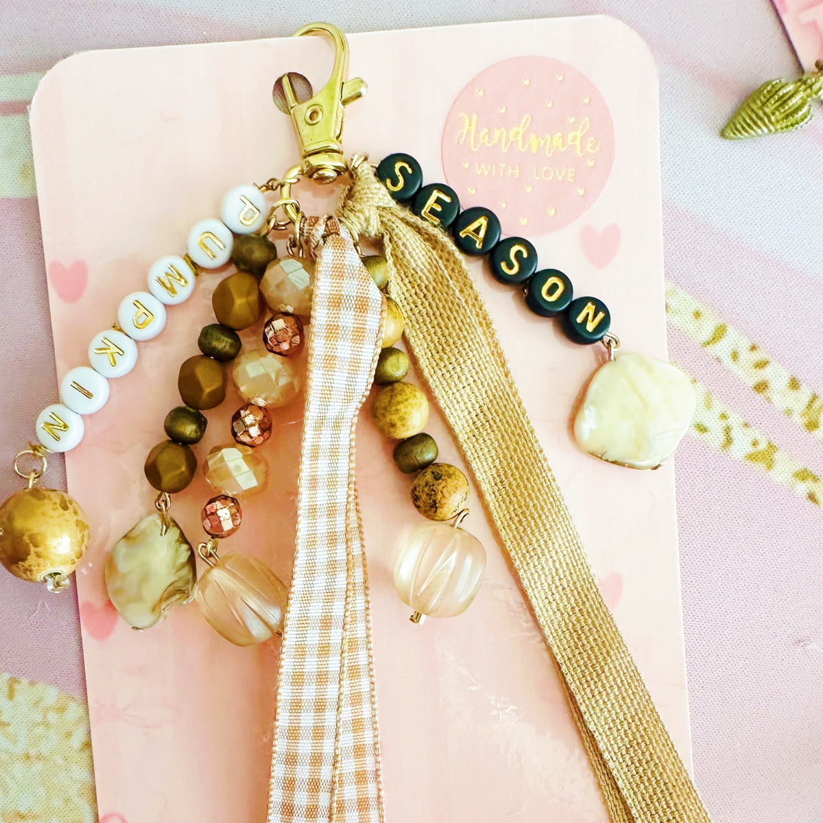 A wbohemian-style beaded keychain charm with wooden beads, clear glass beads, a gold clasp, a 'SEASON' bead string, and two hanging ribbons, displayed on a dark wood background.