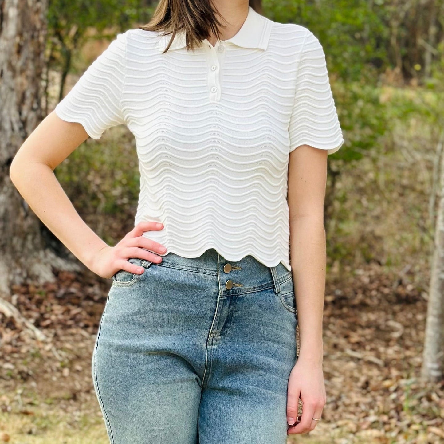 Woman wearing a white top and blue jeans standing in a grassy area with trees.