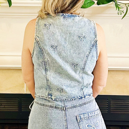 A woman viewed from behind wearing a light-wash denim sleeveless top and matching denim shorts, standing in front of a fireplace in a home setting.