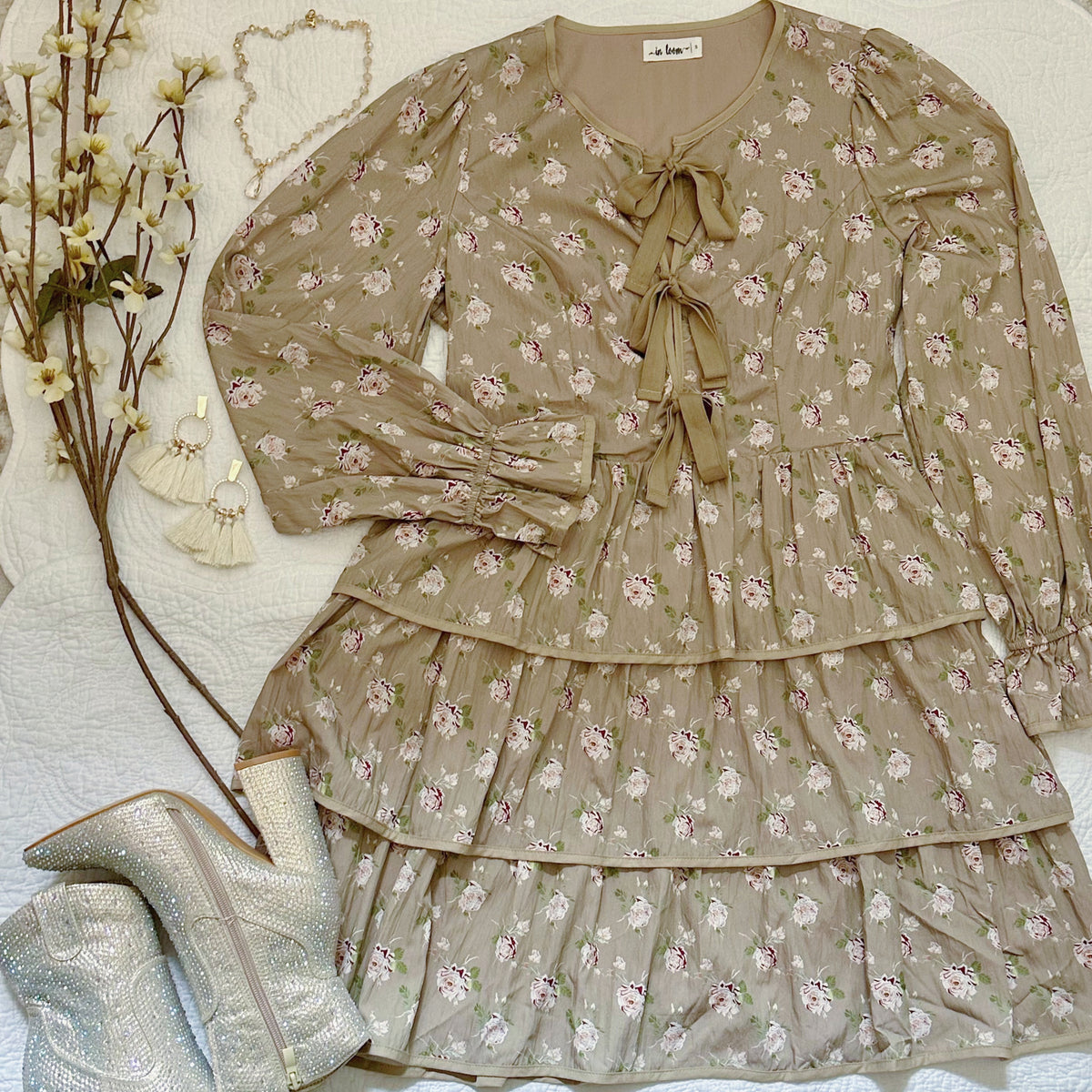 A beige tiered long-sleeve dress with a pink and green floral print, featuring a tie-front neckline and ruffled sleeves, styled in a flat lay with silver glitter ankle boots, cream tassel earrings, and dried flowers on a white textured background.