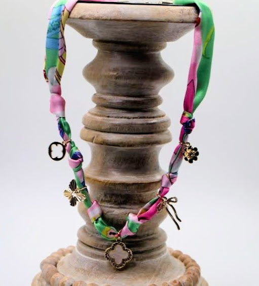 This charming necklace features a vibrant fabric band adorned with intricate charms, including a mother of pearl quatrefoil, a delicate bow, and a lovely flower.
