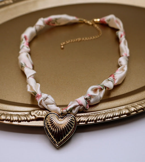 This charming fabric-wrapped choker features a beautifully detailed gold-tone heart charm, elegantly set against a playful floral pattern.