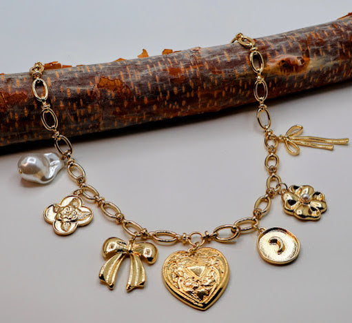 This exquisite necklace features a delightful array of charms, including a metal heart, a charming bow, a floral quatrefoil, and lustrous pearl accents.