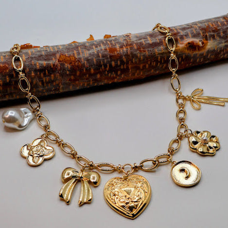 This exquisite necklace features a delightful array of charms, including a metal heart, a charming bow, a floral quatrefoil, and lustrous pearl accents.