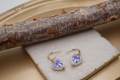 Heart-shaped earrings with blue and white designs on a white stone surface, with a wooden log in the background.