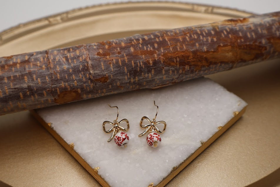 Gold bow-shaped earrings with pink centers on a white stone surface, with a wooden stick in the background.