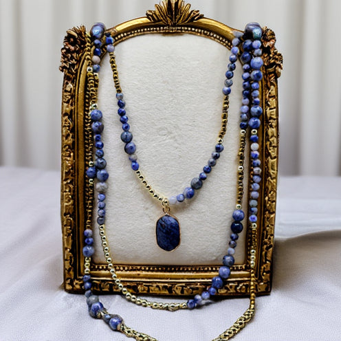 Blue beaded necklace with a gold chain and pendant on a decorative stand.