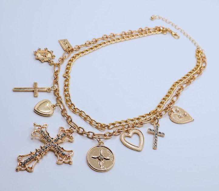 Charm Necklace. This beautifully layered gold-tone necklace features an enchanting collection of delicate charms, including graceful hearts, intricate crosses, and classic coin pendants. The centerpiece, a stunningly detailed cross.