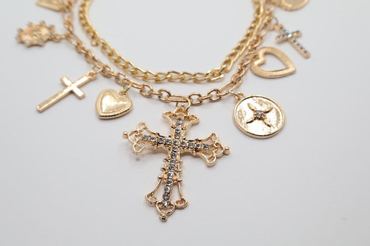 Charm Necklace. This beautifully layered gold-tone necklace features an enchanting collection of delicate charms, including graceful hearts, intricate crosses, and classic coin pendants. The centerpiece, a stunningly detailed cross.