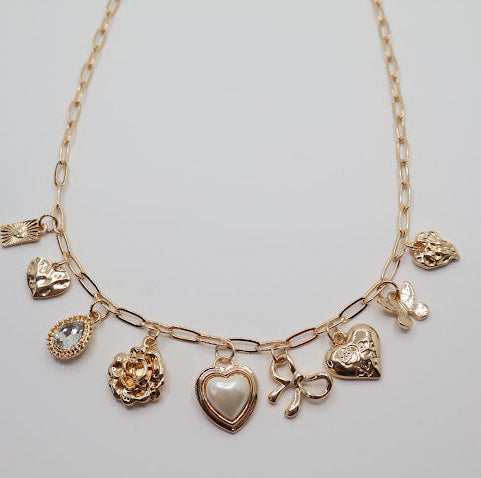 Charm Necklace. Featuring a delicate gold-tone chain adorned with an array of feminine charms, including a pearl heart, a dainty bow, sparkling crystals, and floral motifs.