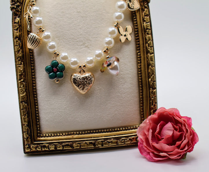 Pearl Charm Necklace is a graceful blend of timeless elegance and whimsical charm. Featuring a strand of lustrous pearls adorned with delicate gold-tone charms, including hearts, flowers, and a touch of sparkle.