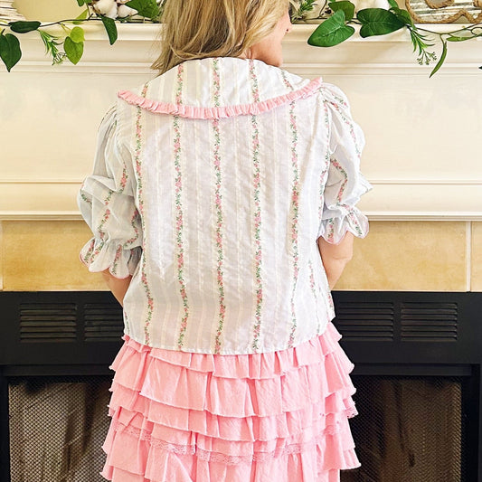 A woman stands with her back to the camera in front of a fireplace, wearing a white ruffled blouse and a pink tiered ruffle skirt, paired with white cowboy boots.