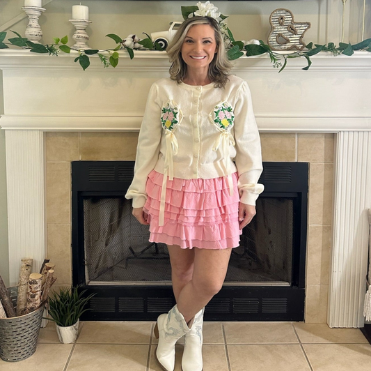Woman in a white blouse with floral designs and a pink skirt standing in front of a fireplace.