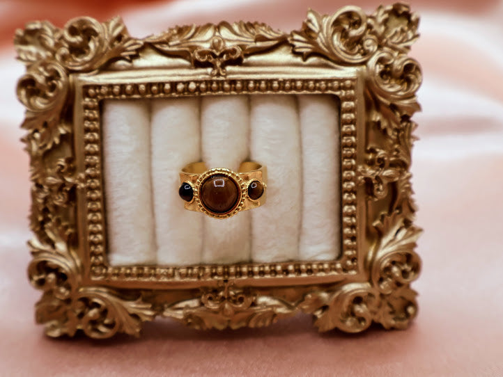 Ring is a captivating blend of warmth and mystique, making it a standout addition to any jewelry collection. Featuring a luminous round amber-toned centerpiece flanked by two smaller dark accent stones.
