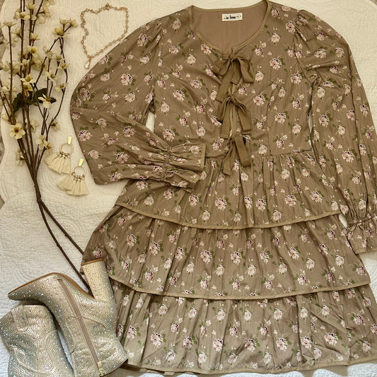 A taupe-brown tiered long-sleeve blouse with a small white floral print, featuring a tie-neck bow and ruffled layers, styled flat-lay with silver glitter ankle boots and dried flowers on a white textured blanket.