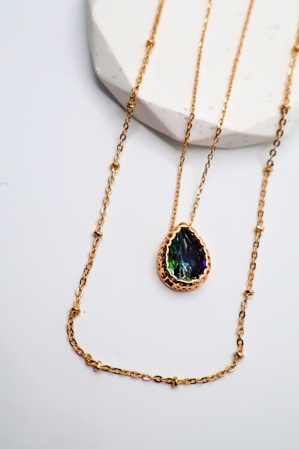 This enchanting layered piece features a stunning teardrop pendant that sparkles with vibrant colors, elegantly suspended between delicate gold chains.