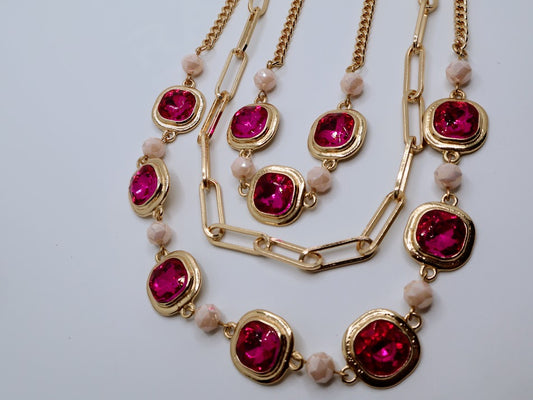 A layered necklace, featuring vibrant ruby-red gems, gold-tone chains, and marbled bead accents.