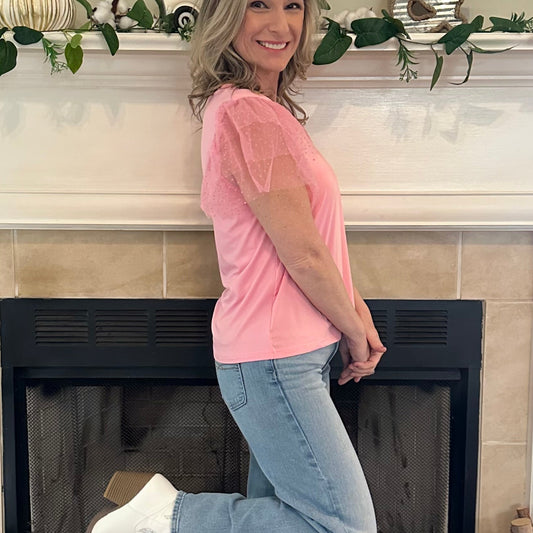 Woman in pink shirt and jeans standing in front of a fireplace with decorative items on the mantel.