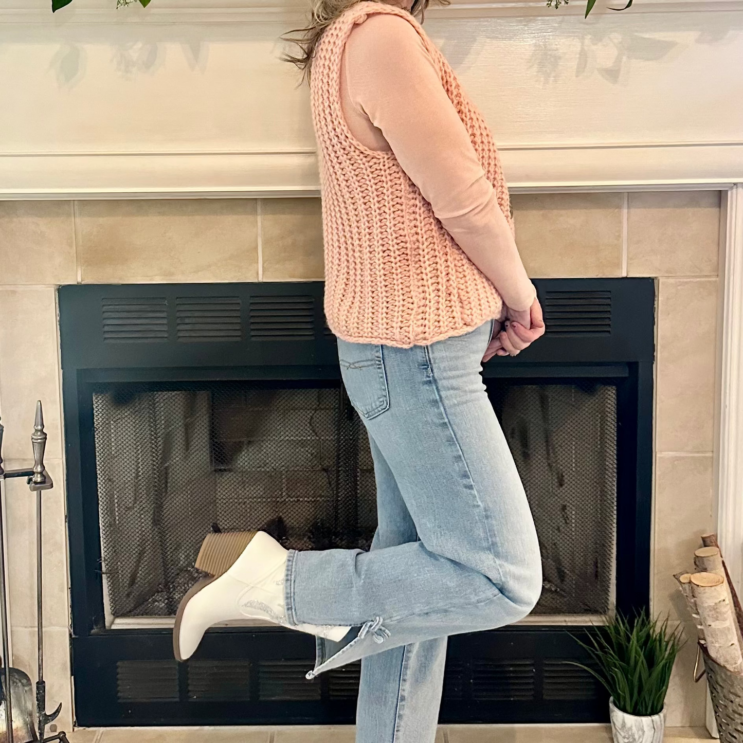 Woman in a pink sweater and jeans standing in front of a fireplace with decorative items on the mantel.