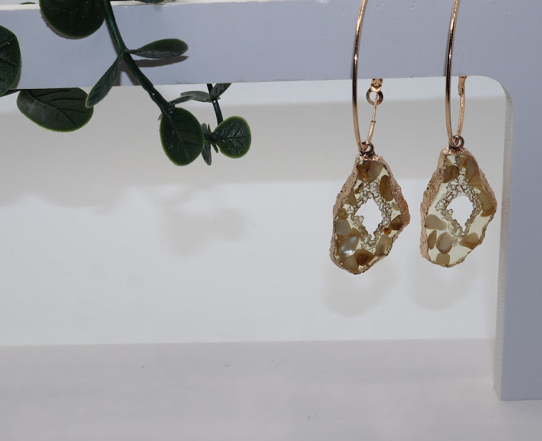 Gold earrings with a natural stone design on a white surface with green leaves in the background