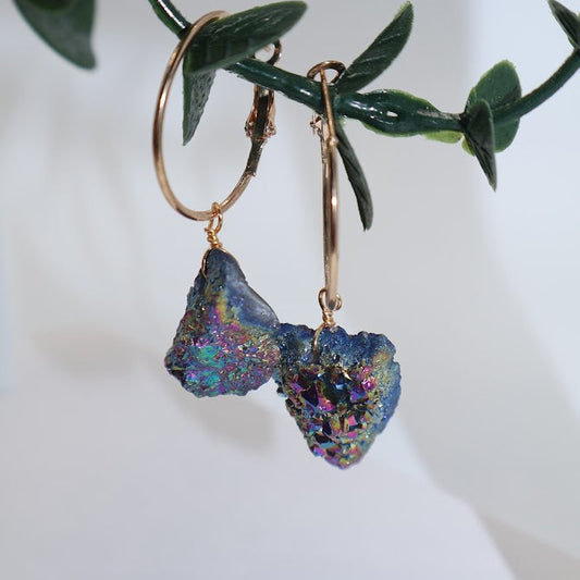 Gold hoop earrings with blue, multicolored stone drops on a white surface with green leaves.