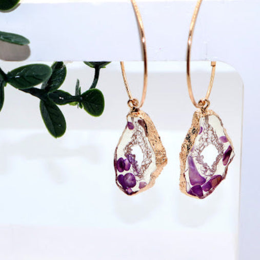 Gold earrings with purple gemstones on a white card with green leaves in the background