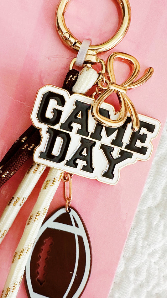 A close-up of a gold-tone keychain with a round key ring, a bow charm, a 'GAME DAY' text charm, and a football-shaped charm, set against a pink background.