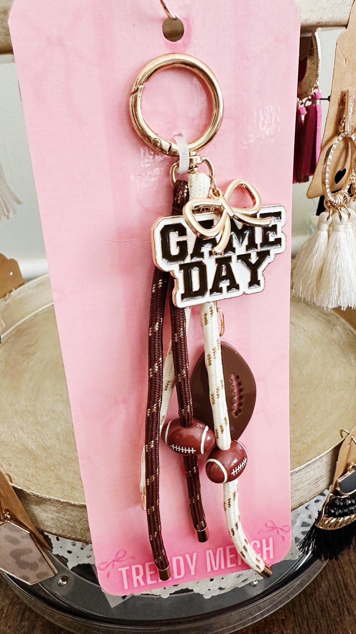 A 'Game Day' themed keychain accessory set on a pink display card. The set includes a gold-tone split ring, a white and black 'GAME DAY' charm, brown and white braided tassels, a brown football-shaped bead, and a small brown football charm.