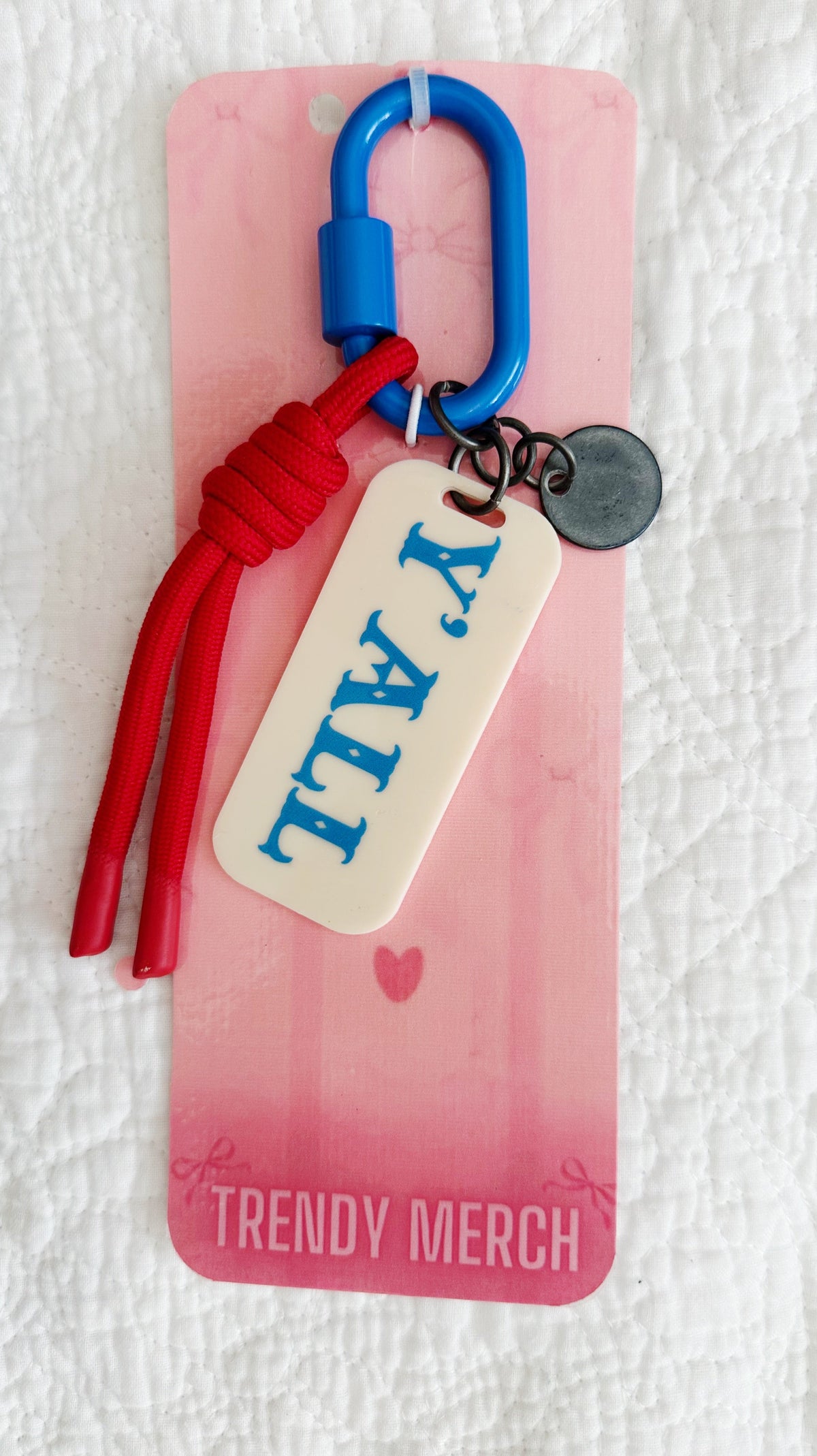 A 'Y'all' keychain charm with a cream-colored rectangular tag, blue carabiner, red cord, and a small round metal tag, attached to a pink 'Trendy Merch' display card on a white textured fabric background.