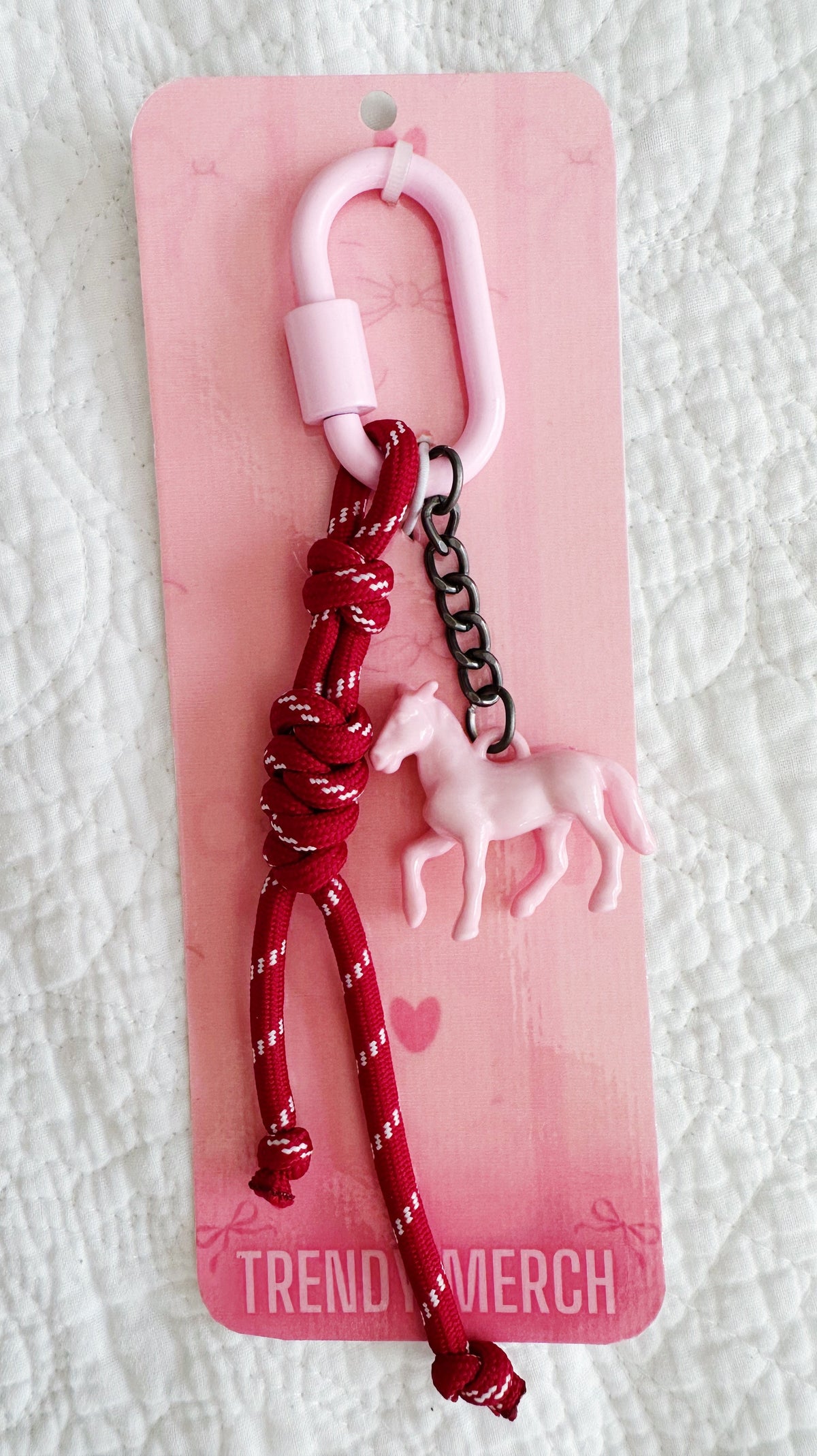 A pink and red keychain or bag charm with a pink horse charm, red braided cord, and black chain, attached to a pink retail card labeled 'TREND MERCH'.