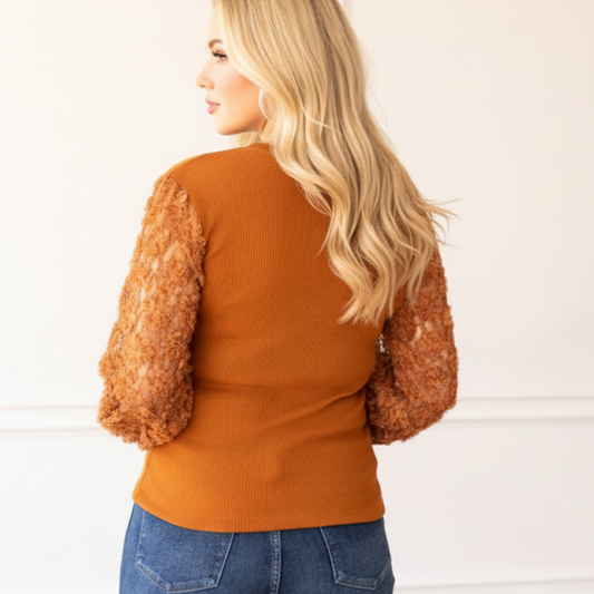 Woman wearing a rust-colored sweater with fur sleeves against a white paneled wall.