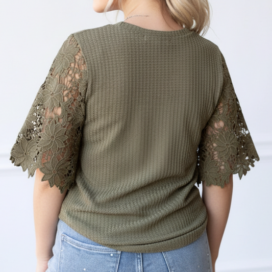 Woman wearing a green top with lace sleeves and light blue jeans against a white wall.