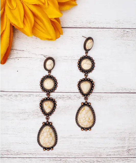 Pair of long, ornate earrings with beige stones on a white wooden surface with yellow flowers.