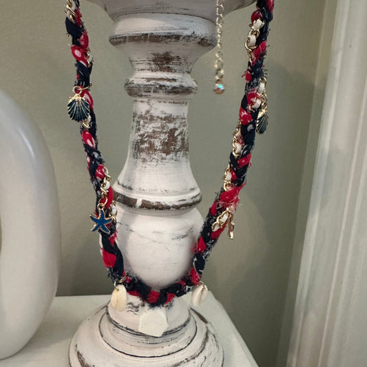 A bohemian-style braided necklace with red, navy, and white braid, decorated with seashells, starfish charms, and a blue star pendant, draped over a distressed white wooden candle holder.