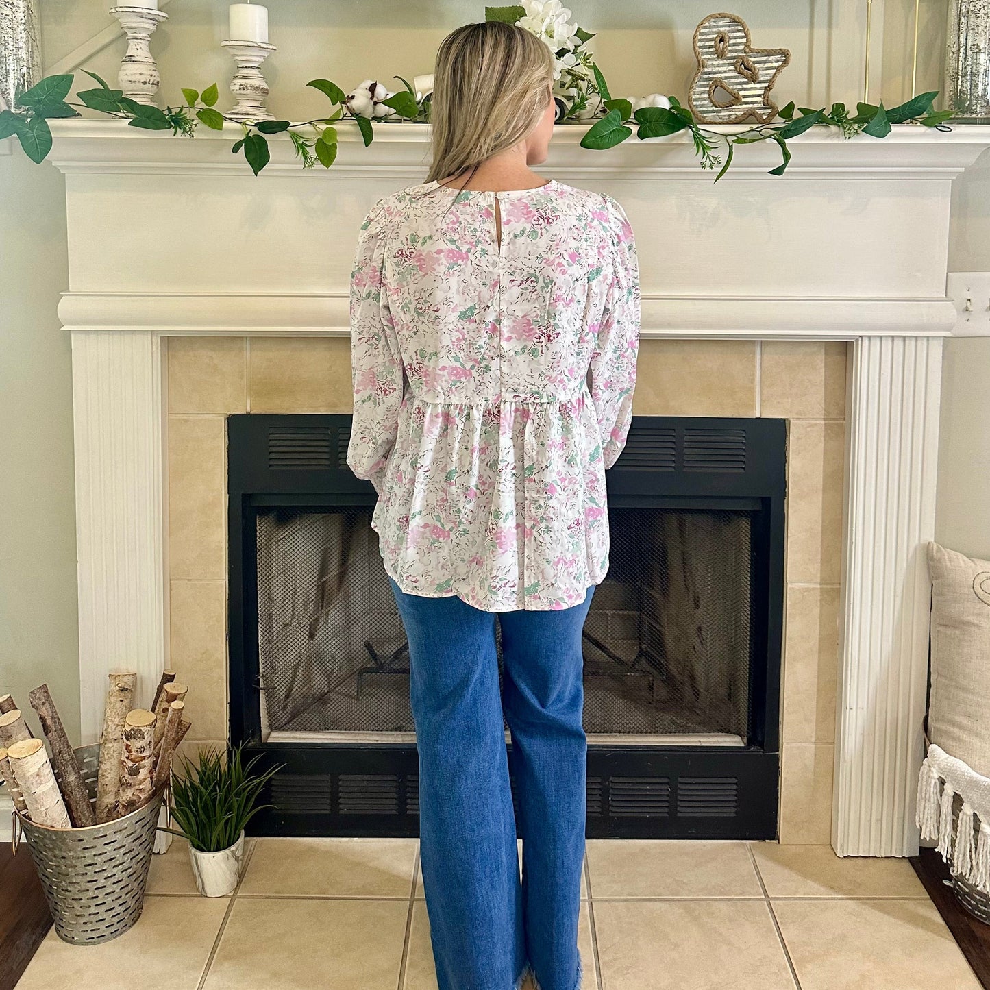 Woman wearing a floral blouse and blue jeans standing in front of a fireplace.