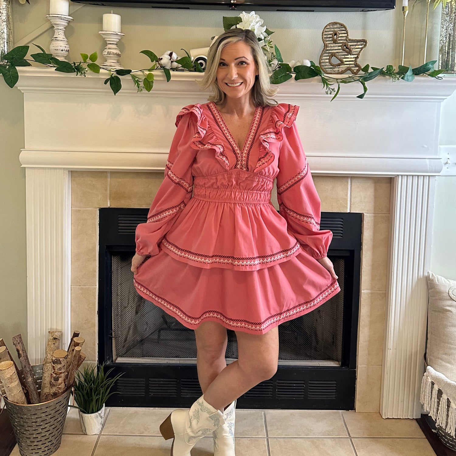 Woman wearing a pink dress with ruffled sleeves and trim in front of a fireplace.