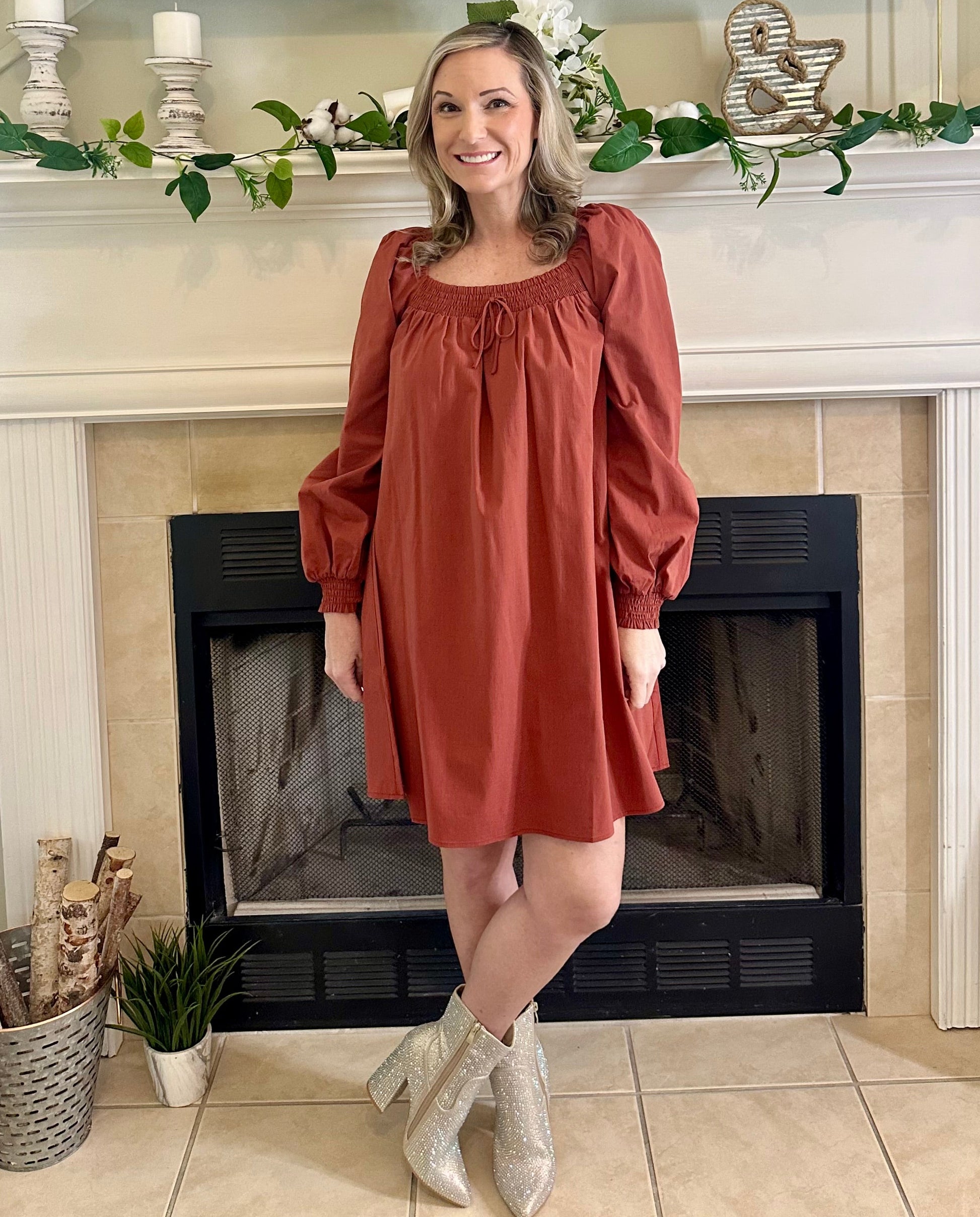 Woman in a rust-colored dress standing in front of a fireplace with decorative elements.
