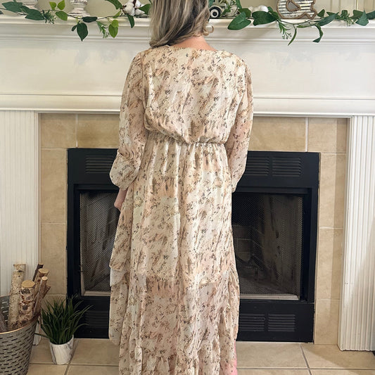 A woman stands with her back to the camera wearing a long-sleeved, tiered maxi dress with a beige and pink floral pattern, positioned in front of a fireplace.