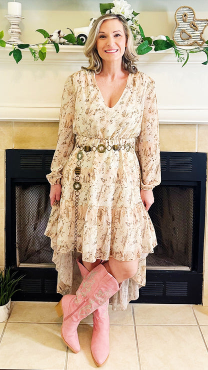 A woman stands in front of a fireplace wearing a long-sleeve, tiered cream floral dress with a decorative belt and pink cowboy boots.