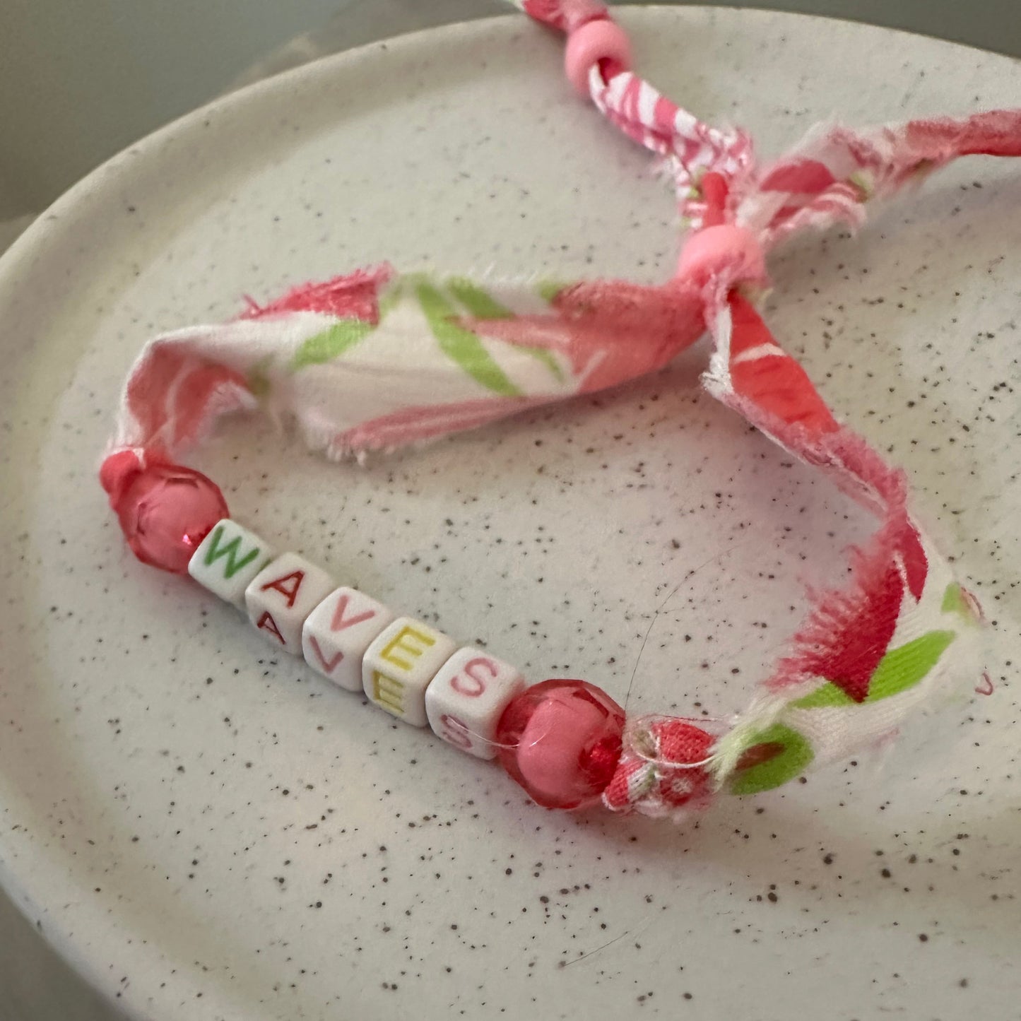 A handmade fabric and bead bracelet with pink and white striped frayed cords, pink beads, and white letter beads spelling 'WAVE'S', displayed on a speckled white ceramic plate.