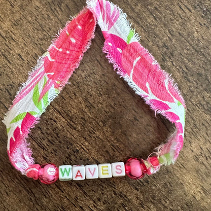 A handmade pink fabric bracelet with a floral print, frayed edges, and pink beads, featuring a letter bead spelling 'WAVES' on a dark wood background.