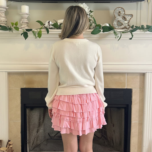 Person wearing a pink ruffled skirt and white sweater standing in front of a fireplace.